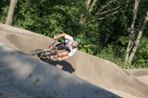 A man in a white shirt and black shorts rides a BMX bike on a dirt track, surrounded by lush green trees.
