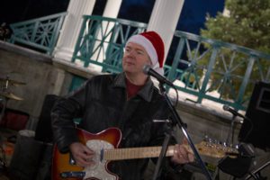 A man in a Santa hat and leather jacket plays an electric guitar at an outdoor venue, with a microphone and festive backdrop.