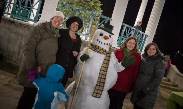 Four women and a child pose with a snowman wearing a hat and scarf in front of a gazebo with columns and a railing.