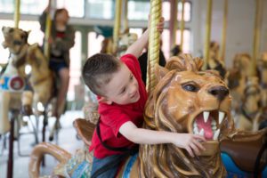 A young boy in a red shirt joyfully rides a carousel lion, gripping the pole and reaching for the lion's mouth.
