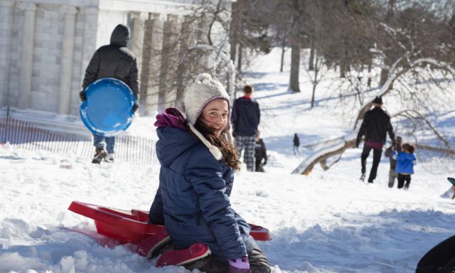 A girl in a blue coat and white hat smiles while sitting on a red sled in the snow. Others are sledding nearby, with a large building in the background.