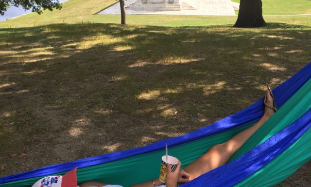 A person relaxes in a blue and green hammock, wearing a Pabst Blue Ribbon cap, holding a drink. A monument and lake are in the background.