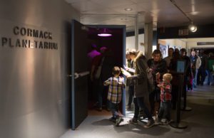 Visitors enter Cormack Planetarium through a dimly lit door, with a crowd waiting in line outside.