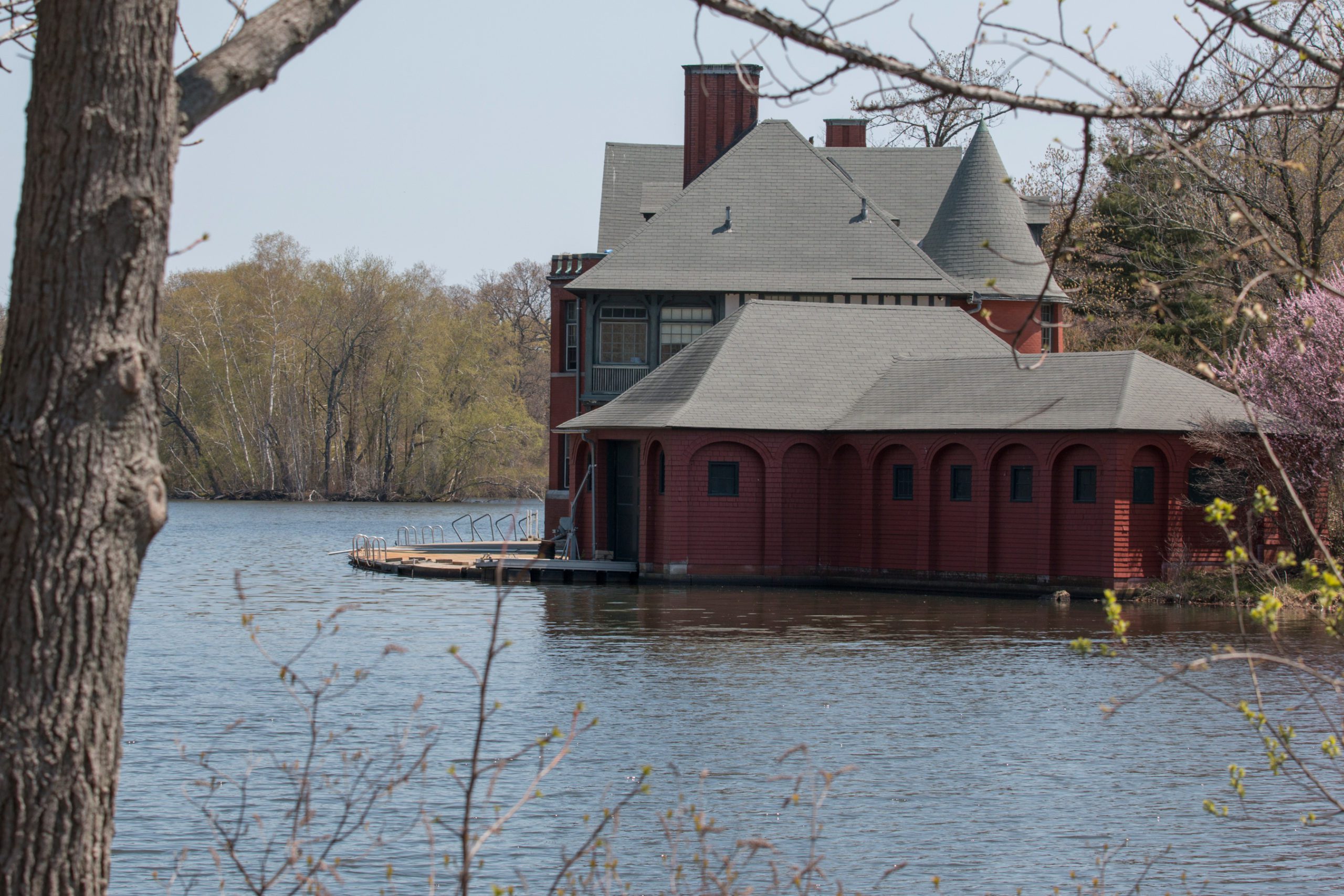 A red boathouse with arched windows sits by the water's edge, surrounded by trees and foliage on a clear day.