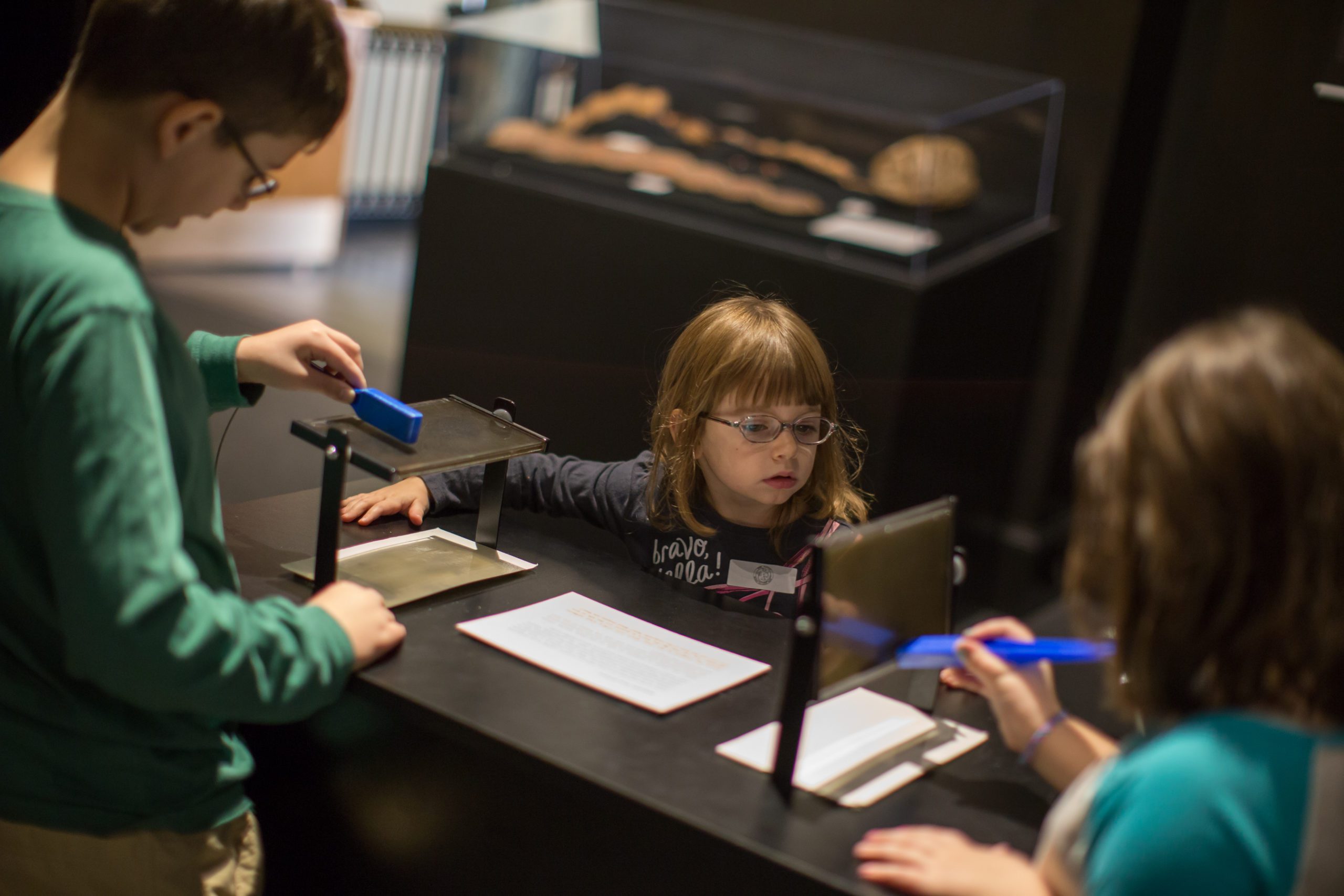Children interacting with an exhibit at a museum, using magnifying tools to examine objects on a table.