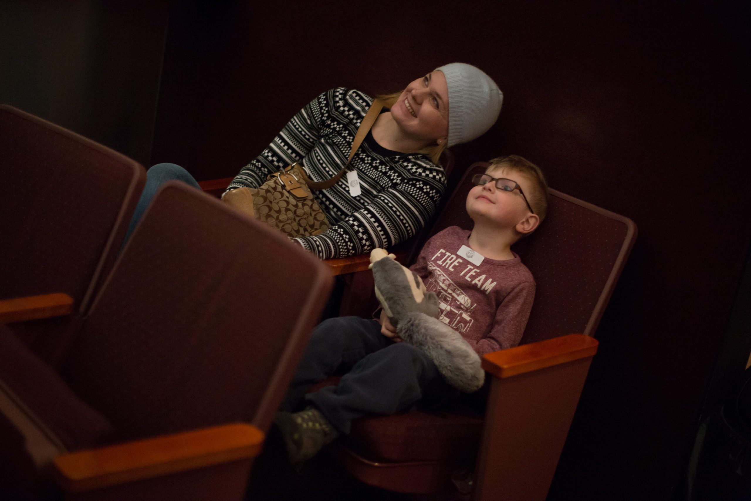 A woman wearing a winter hat and a patterned sweater sits in a theater seat, smiling. Next to her, a boy with glasses holds a stuffed animal, looking content.