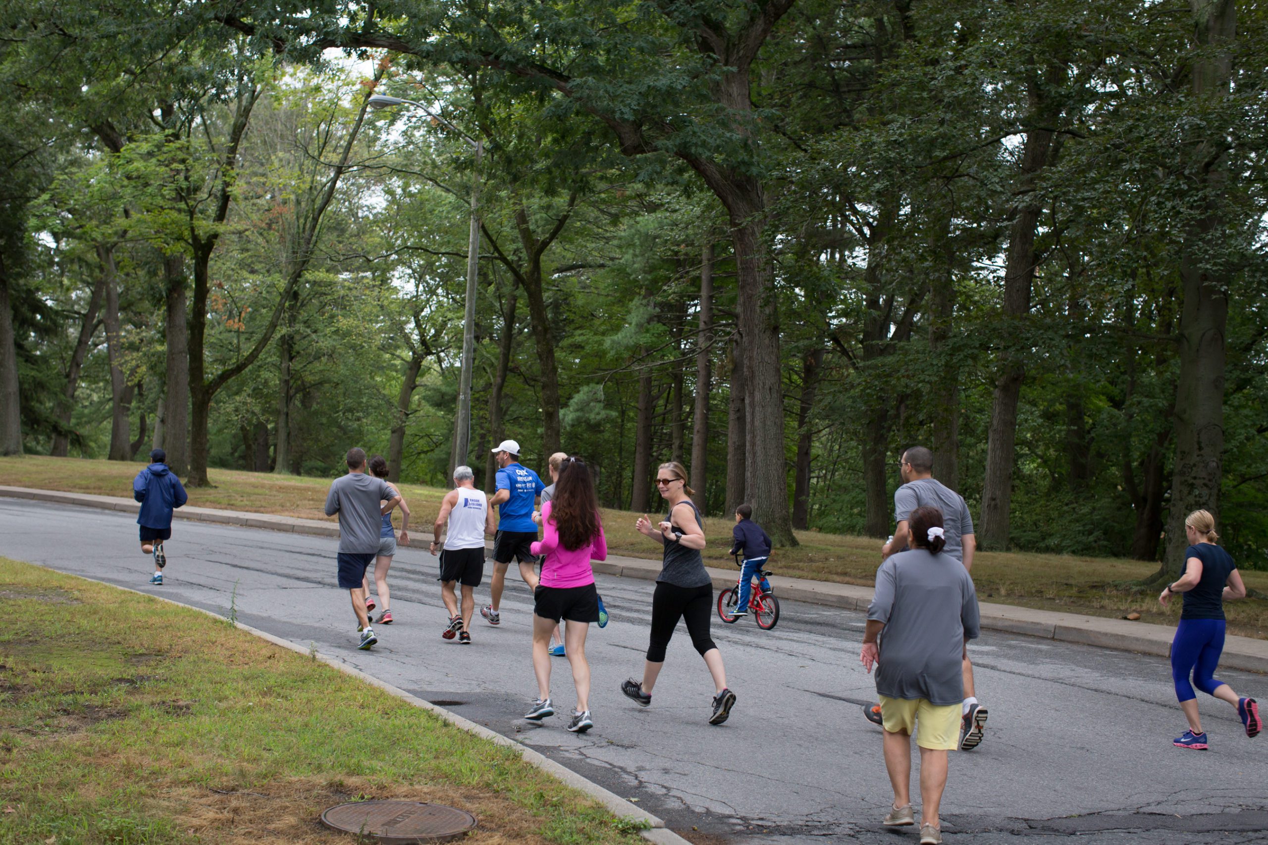 People jogging on a tree-lined park road, with a child on a red bike. The scene is lively and surrounded by greenery.