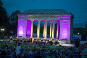 Outdoor concert by the Rhode Island Philharmonic at the Temple to Music, lit in purple and yellow, with a large audience seated on the grass.