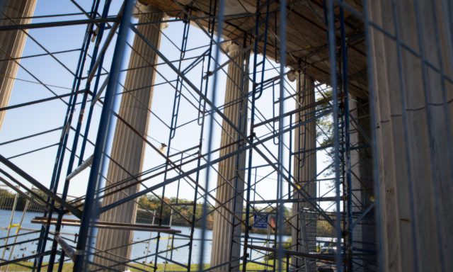 Scaffolding surrounds large columns overlooking a lake, with a clear sky and trees in the background.