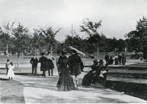 People stroll in a park on a sunny day, with women in long dresses and a person holding a parasol. Trees and pathways are visible.