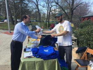 A man in a blue shirt exchanges a plastic bag for a reusable one with another man in a park setting during Earth Day.