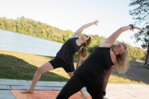 Two women practice yoga outdoors by a lake, stretching with one arm raised. The scene is peaceful with trees and clear skies.