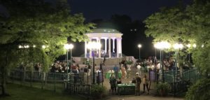 Nighttime event at Roger Williams Park, with a lit pavilion and people mingling around tables under trees and streetlights.