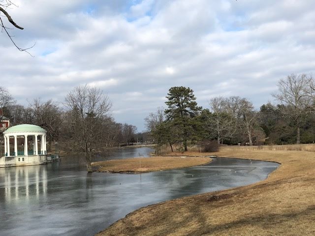 A serene, late afternoon scene at Maymont Park featuring a frozen pond, a small island, trees, and a gazebo under a cloudy sky.