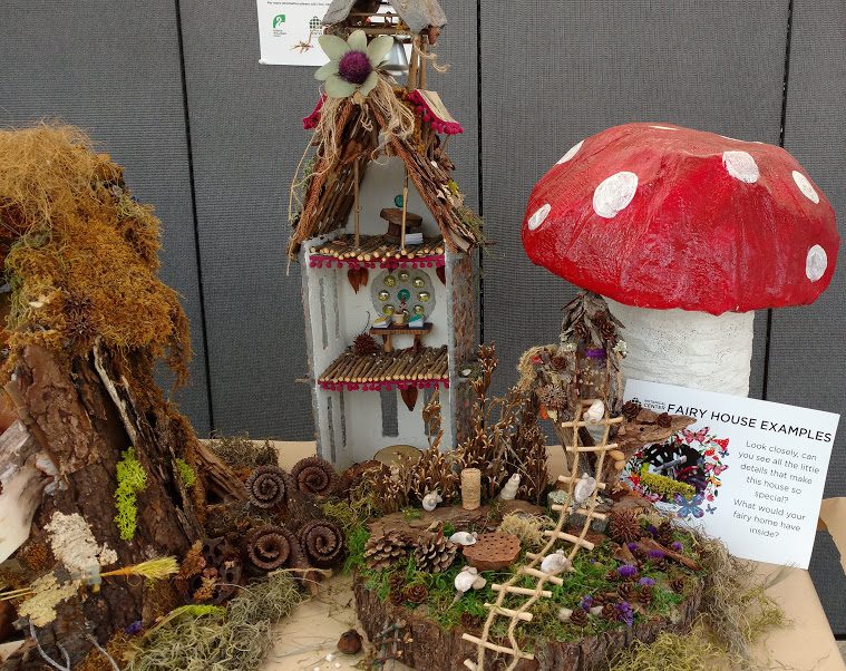 Fairy house display featuring a multi-level structure with natural elements, a red mushroom with white spots, and a sign titled "Fairy House Examples."