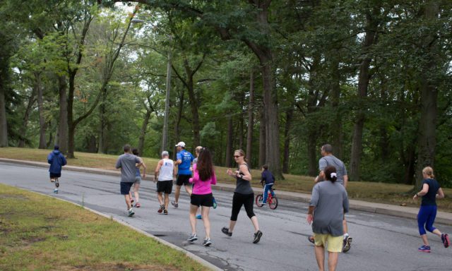 A group of people running and walking on a tree-lined path, with a child on a red bicycle, in a park setting.