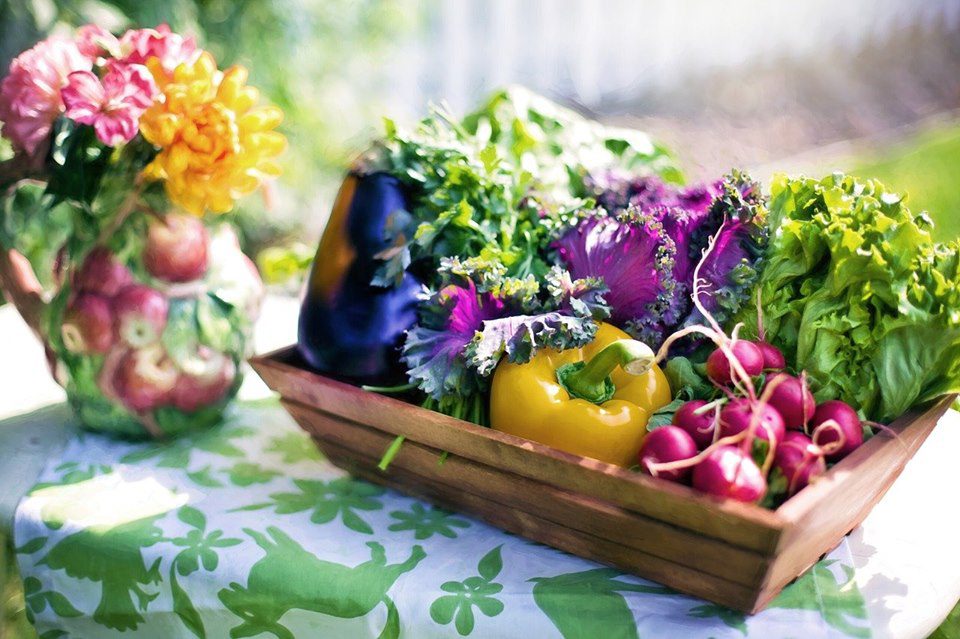 A wooden basket filled with fresh vegetables, including a yellow bell pepper, eggplant, radishes, and leafy greens. A colorful floral vase is nearby on a table with a green-patterned cloth.