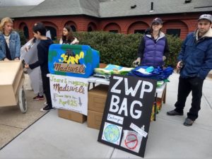 People gather at an outdoor booth with signs promoting Madewell's jeans and magazine collection and a Zero Waste Program bag exchange.