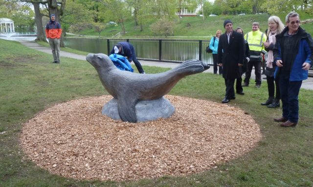 A group of people gather around a sea lion statue in a park. One person adjusts a blue cloth on the statue.