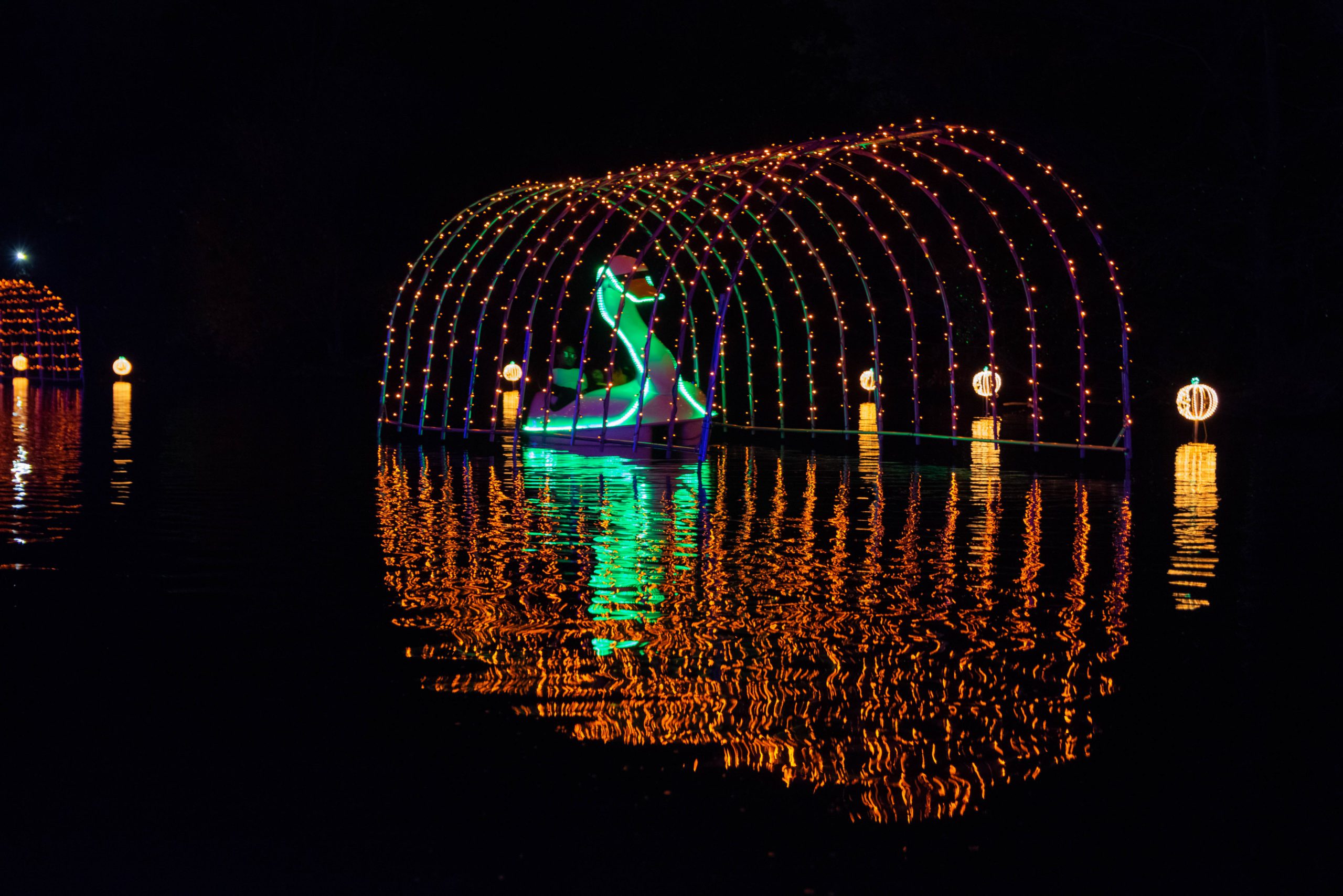 A swan boat illuminated with vibrant lights under an arch of colorful bulbs reflects on the water at night.