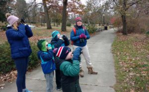A group of people, including children, are birdwatching in a park. They are using binoculars and dressed warmly for the cool weather.