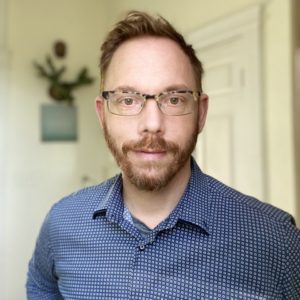 Man with short light brown hair, a trimmed beard, and tortoiseshell glasses standing indoors. He is wearing a blue patterned button-up shirt and looking directly at the camera with a neutral expression. The background shows a softly lit room with white walls and a blurred plant and artwork.