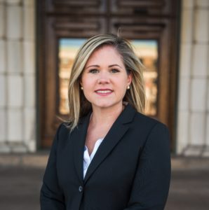 Smiling woman with blonde hair in a black blazer, standing in front of a wooden door.