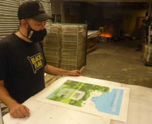 A man in a Black Flag shirt and cap, wearing a mask, examines a Roger Williams Park print in a workshop.
