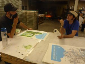Two men discuss prints at a workshop table. One wears a mask and cap, the other holds paper towels. A poster reads "Roger Williams Park."