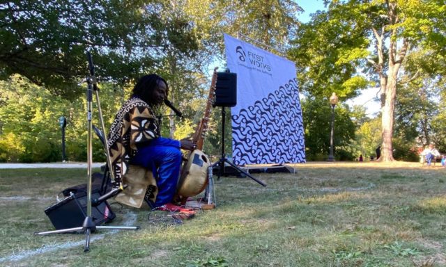Musician performs outdoors with a kora beside a "First Works" banner. Trees and spectators are visible in the background.