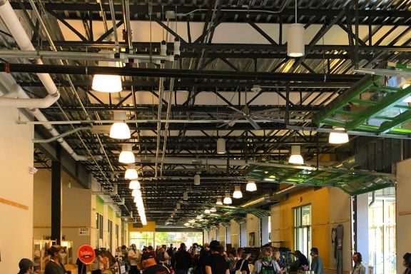 A bustling indoor market with people browsing various stalls selling fresh produce and goods, under a high industrial-style ceiling.