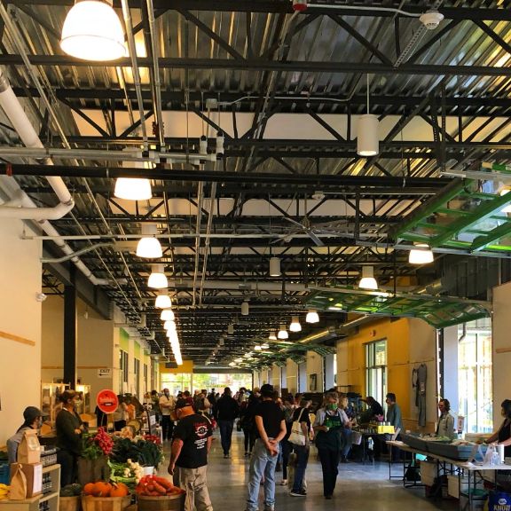 A bustling indoor market with people browsing various stalls selling fresh produce and goods, under a high industrial-style ceiling.