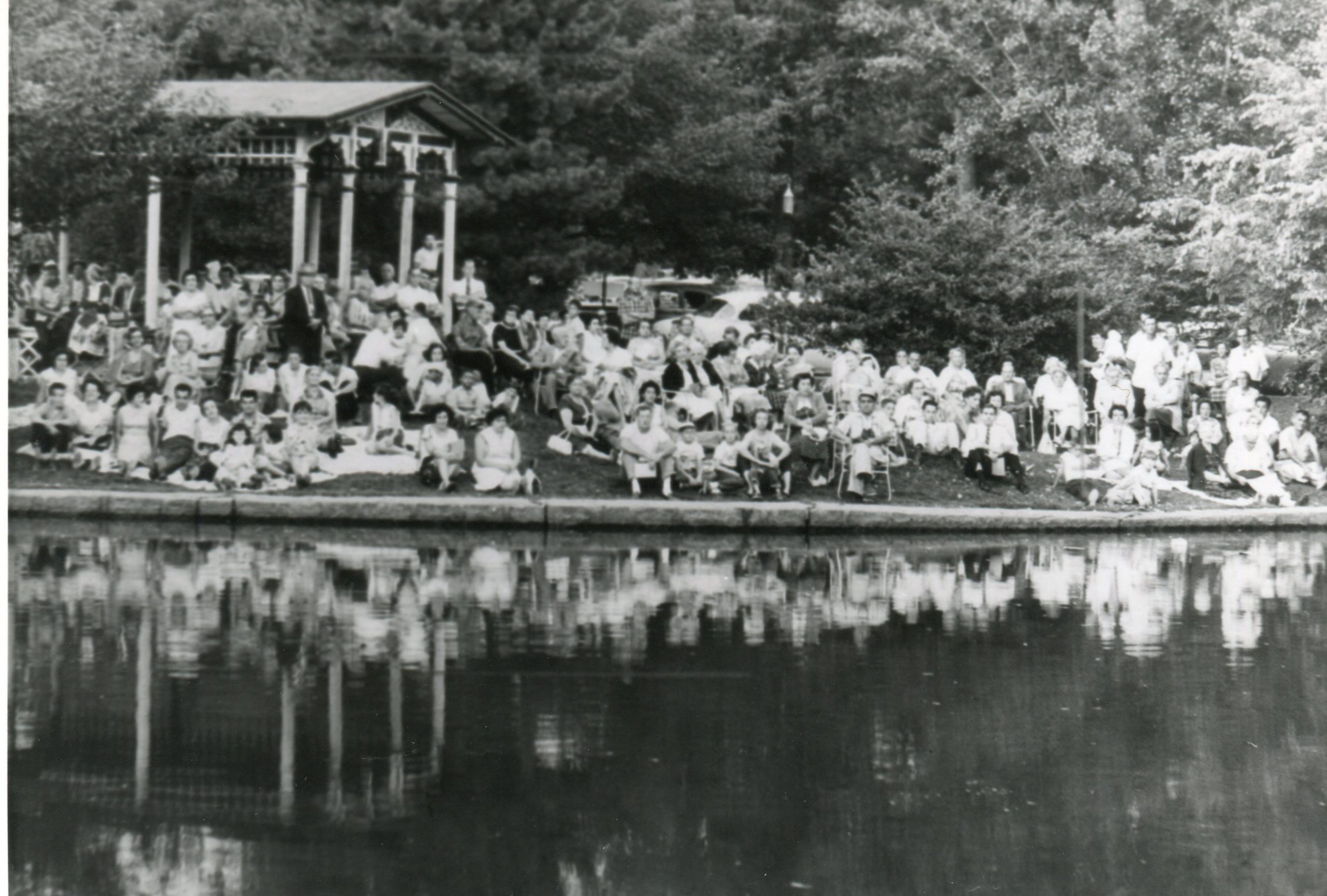 A large group of people gathers near a pond, sitting on the grass and in chairs, with a gazebo in the background surrounded by trees.