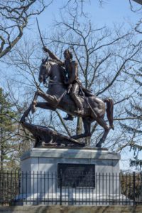 Statue of Casimir Pulaski on horseback, sword raised, in Roger Williams Park, Providence. Surrounded by bare trees and a metal fence.