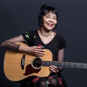 Smiling woman with black hair holds an acoustic guitar, wearing a floral dress against a dark background.