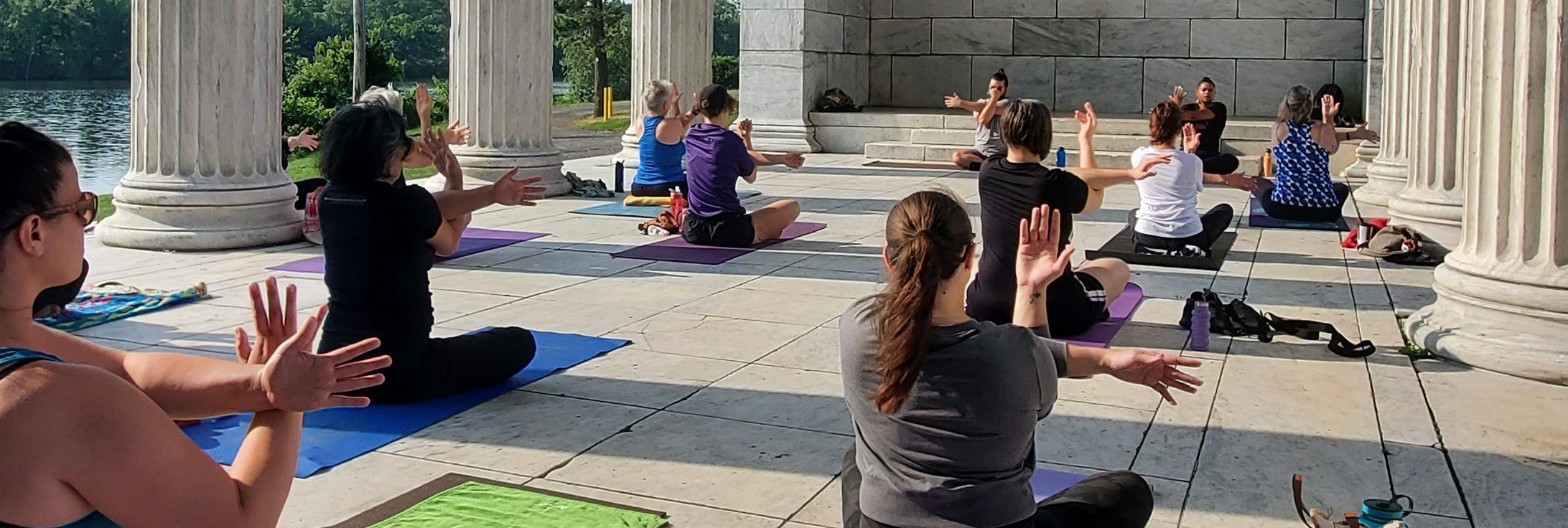 A group of people practicing yoga on mats in an outdoor pavilion with columns, near a lake.