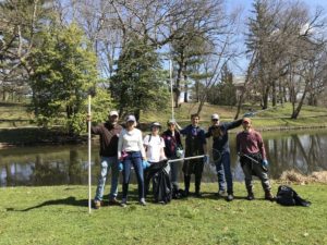 A group of seven people stands by a pond, holding cleaning tools and bags, participating in an Earth Day cleanup event in a park.