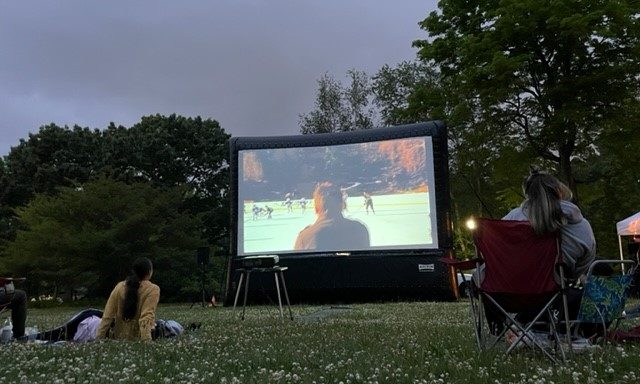 Outdoor movie night at First Friday Film, with people seated on the grass watching a film on a large inflatable screen surrounded by trees.