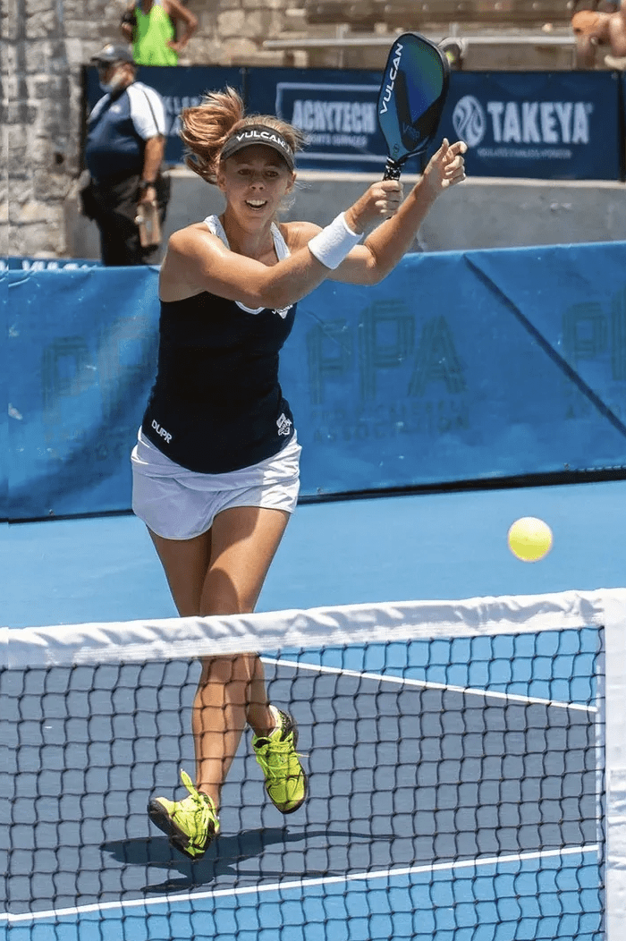 A player in action on a blue pickleball court, wearing a Vulcan visor and holding a Vulcan paddle, with a ball approaching the net.