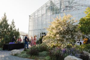 A group of people socialize at a spring event outside a glass conservatory. A table with drinks is set up nearby amidst lush greenery.