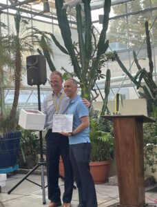 Essington and Elorza at an award event in a botanical center, with Elorza holding a certificate and Essington a box.