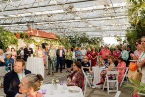 A gathering at a botanical center features attendees in colorful attire, seated and standing, surrounded by lush greenery and a tiled roof structure.