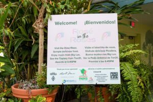 Welcome sign for "Rosé in the Roses" event at a botanical center, featuring activities, sponsors, and bilingual text in English and Spanish.