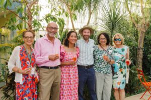 A group of six people smiling and holding drinks, standing in a lush garden setting with vibrant green plants.