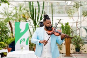 A man in a light blue blazer plays the violin in a botanical center, surrounded by lush plants. A "MusicBotanicalCenter" sign is visible.