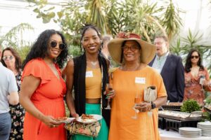 Women at a botanical center party: Vanessa in a mustard top, Eleanor in an orange dress and hat, and another in a red dress.