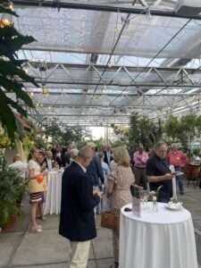 A group of people mingling at a fundraiser event in a greenhouse. Tables with white cloths, drinks, and plants are visible.