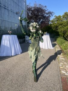 A performer dressed in a floral-themed costume poses outdoors near tables with white tablecloths and flower arrangements.
