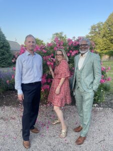 Three people pose in front of vibrant pink roses. The man on the left wears a light shirt and dark pants. The woman in the center wears a red floral dress and glasses, smiling while holding a rose. The man on the right wears a green plaid suit and glasses. The background features a clear blue sky and green trees.