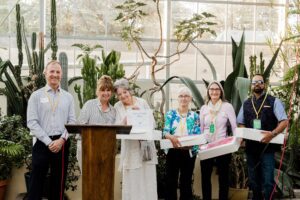 Volunteers at a botanical center receive awards, standing in front of cacti. Kevin and others smile, holding certificates and boxes.